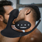 In the background, two men are kissing passionately. In the foreground, there is a black OhMiBod Club Vibe 3.OH Hero Vibrating Anal Plug made from body-safe silicone, with control buttons clearly visible. The device seems to be intended for intimate activities. The setting is a dimly lit room.